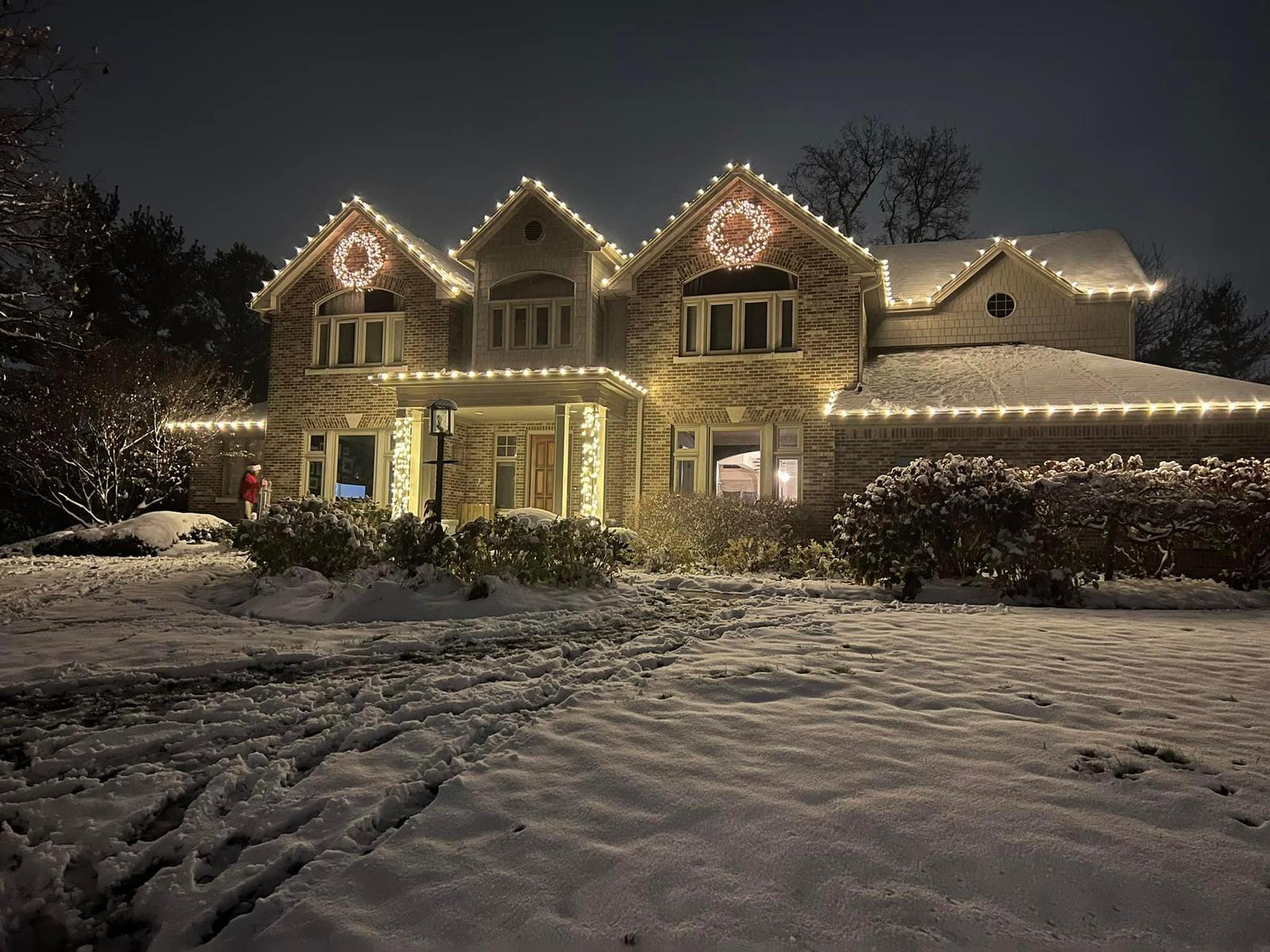 Warm white Christmas lights installed on a brick two-story home in Indiana with lighted wreaths and snow